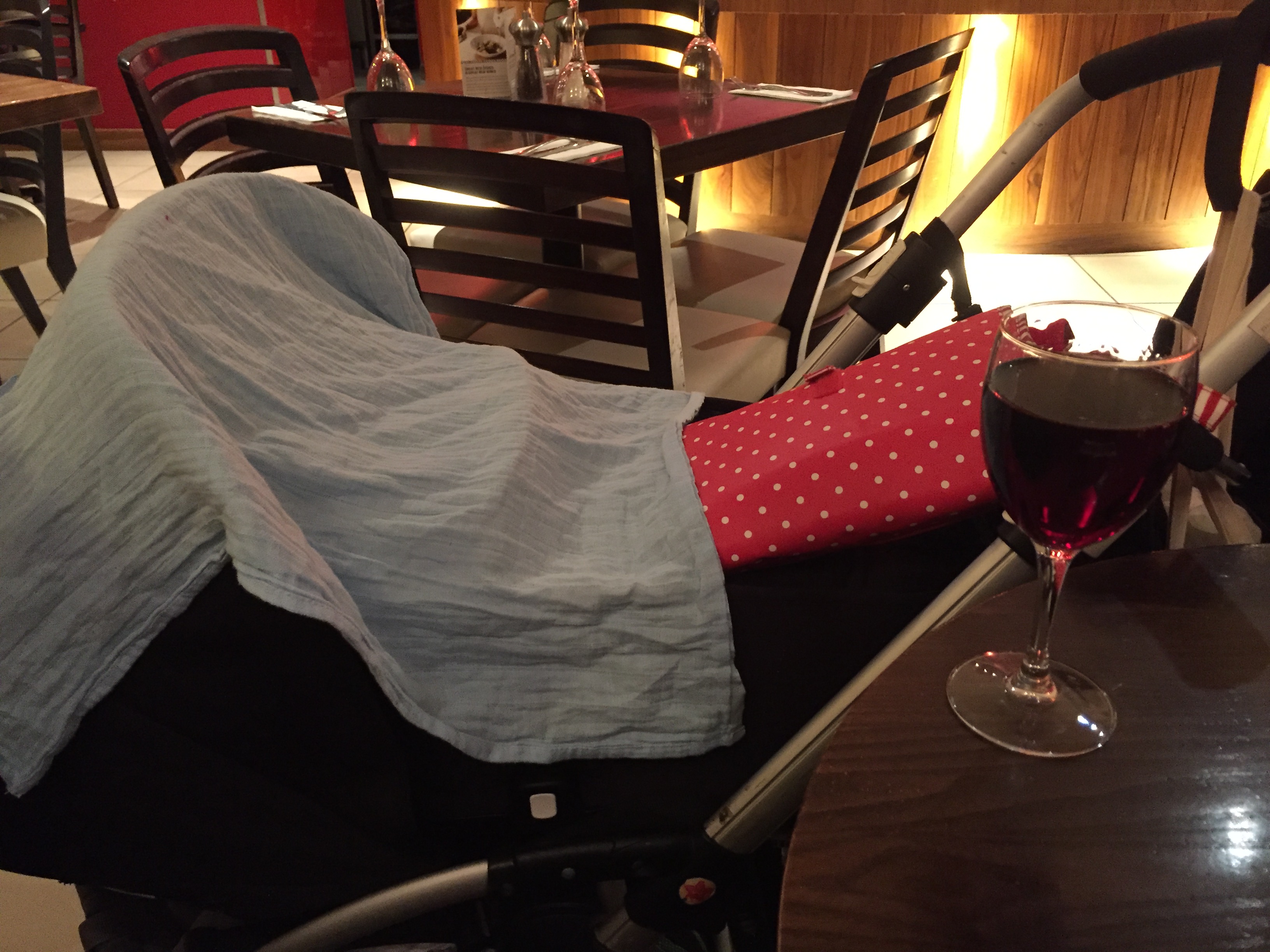A pram with a cloth covering a sleeping baby is parked next to a restaurant table with a glass of red wine on it