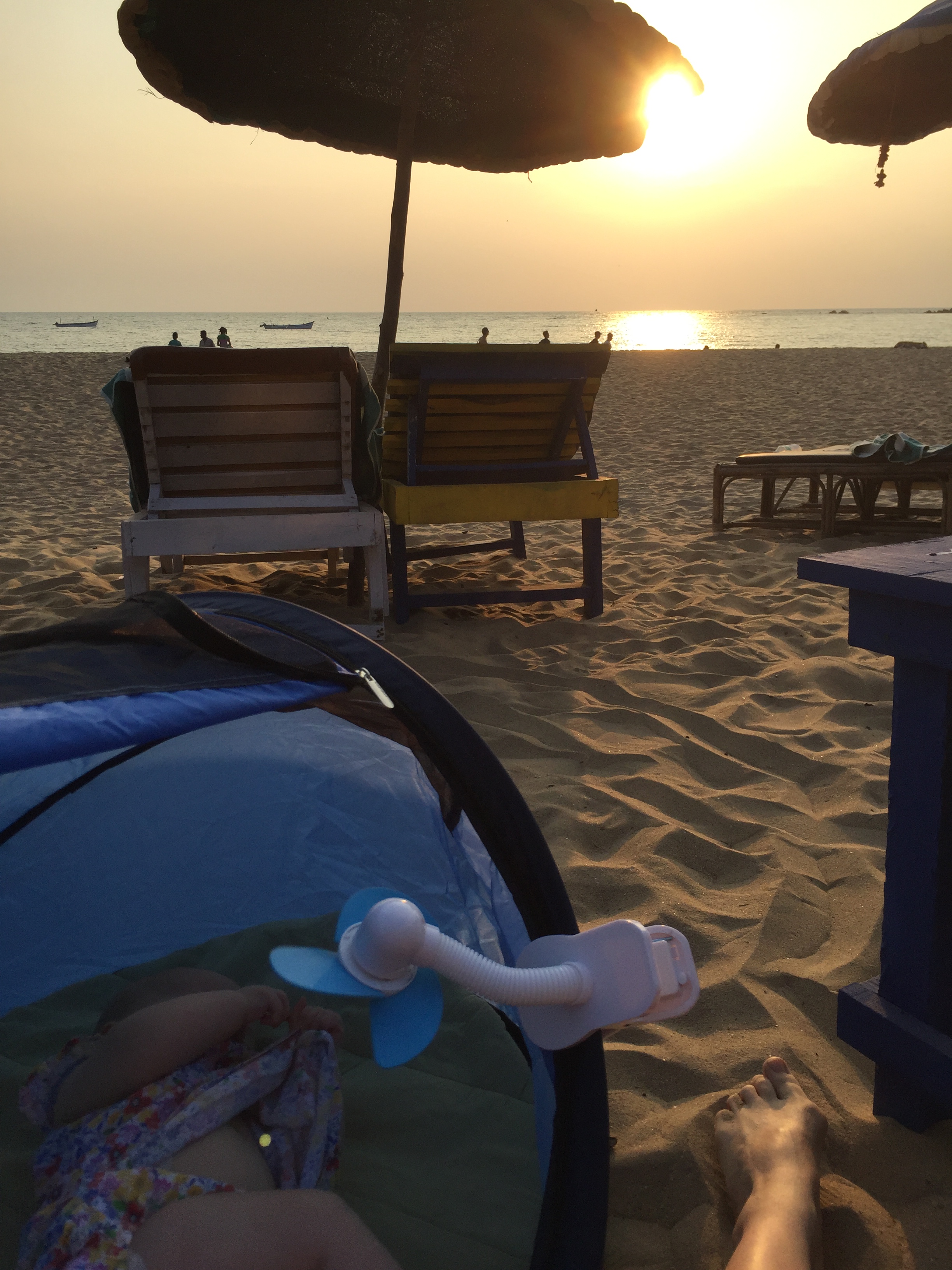 Baby on holiday in a pop-up tent travel cot on a beach in Goa, with the sun setting over the sea. A mini fan is keeping the baby cool. There are sun loungers on the beach.