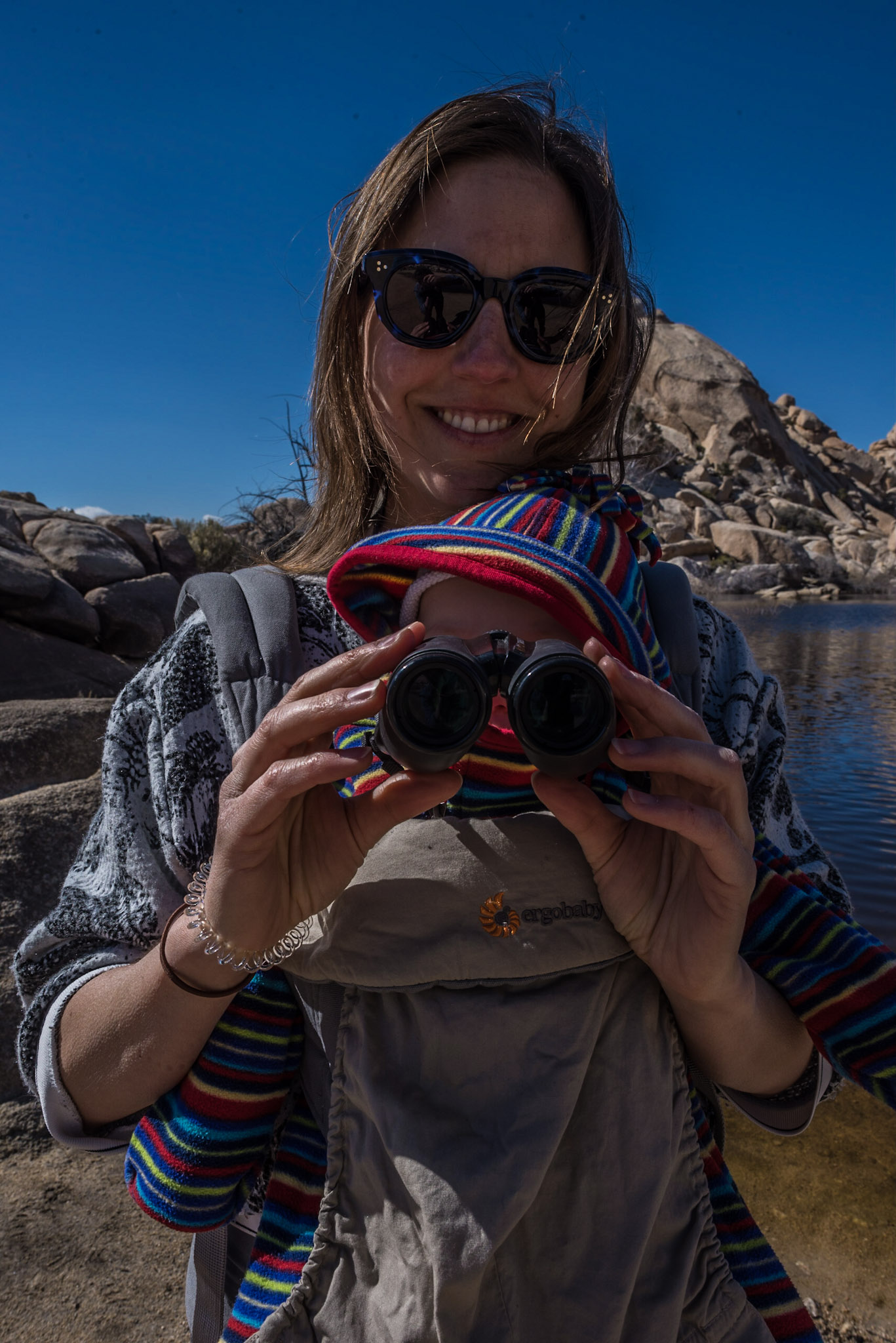 A woman carries a baby in a sling, holding a pair of binoculars up to the baby's face. In the background is the rocky landscape with a lake.