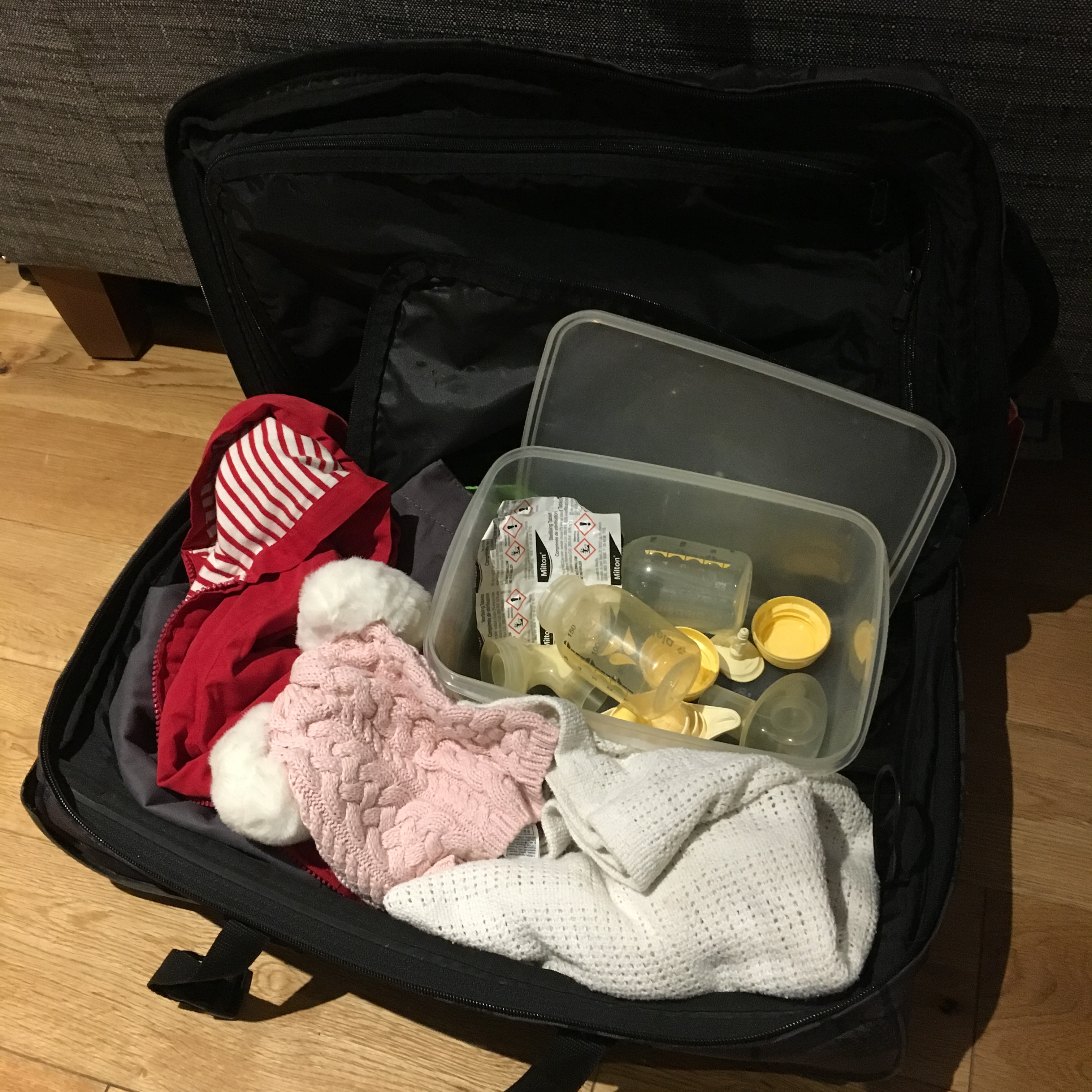Essential kit for cold sterilising – feeding equipment, sterilising tablets, Tupperware box – in a partially packed suitcase filled with baby clothes
