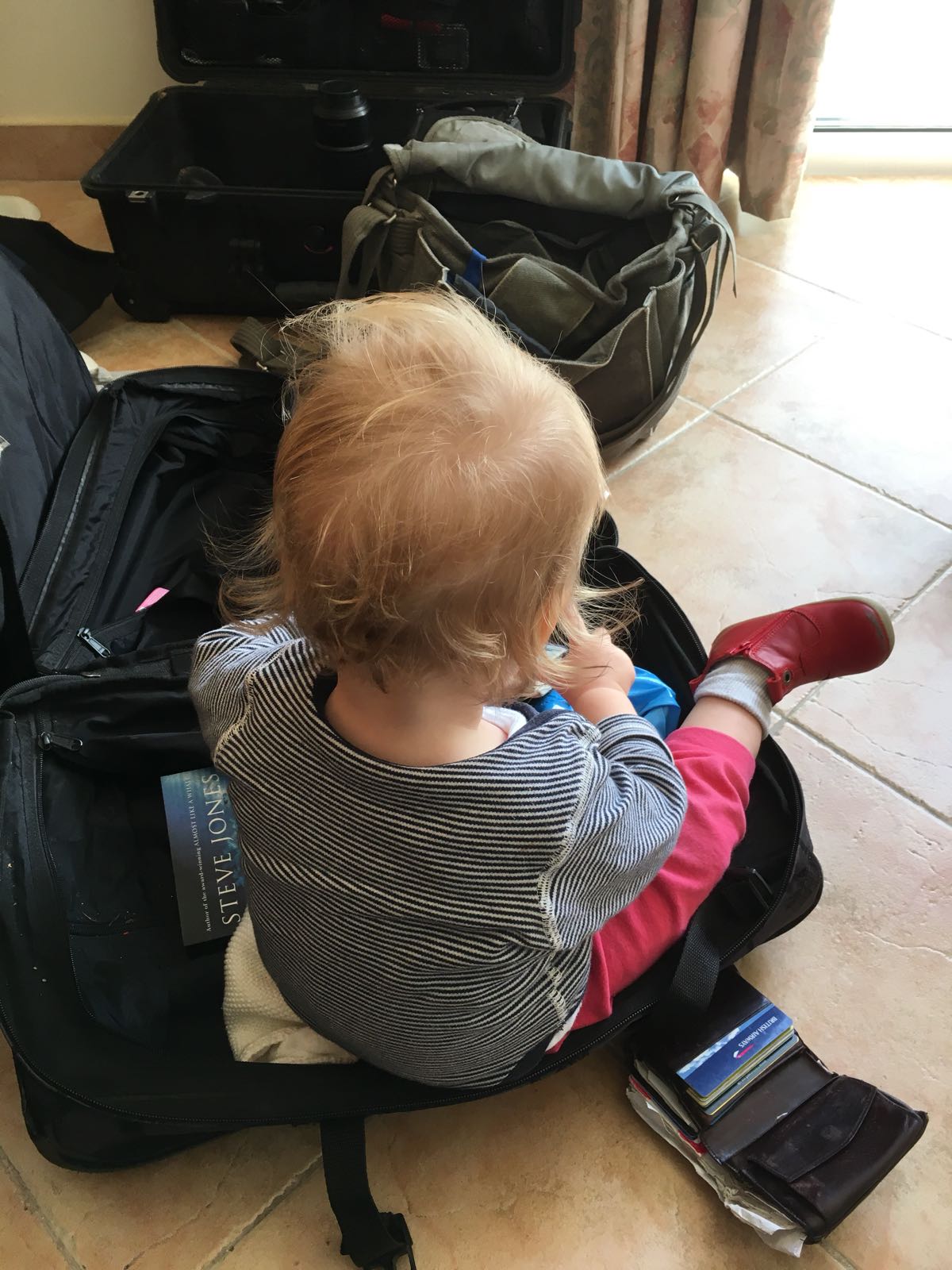 A toddler sits in an open suitcase, other bags on the floor around her.