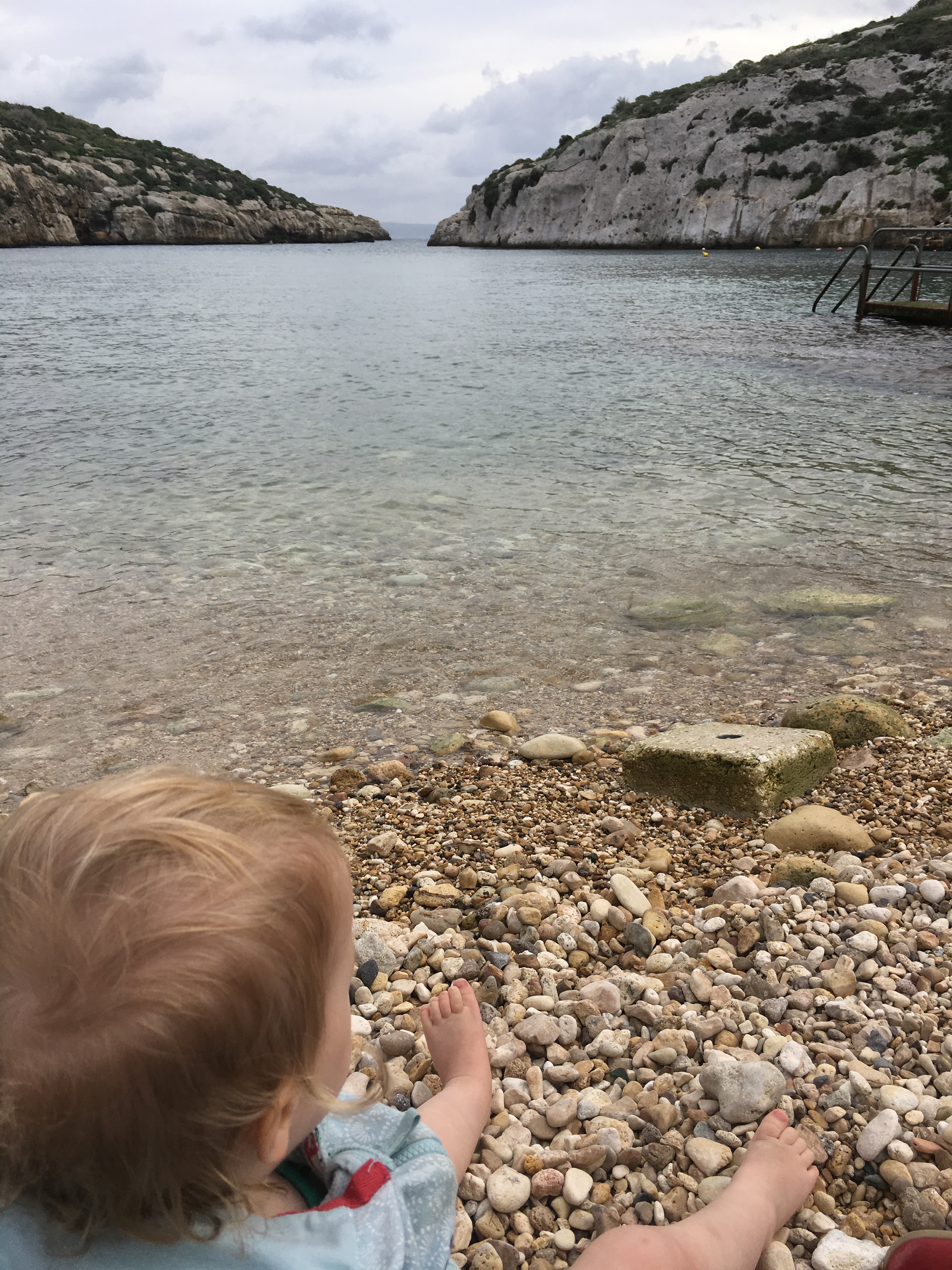 A blonde baby sits on a pebbly beach, looking out over a bay with crystal clear water