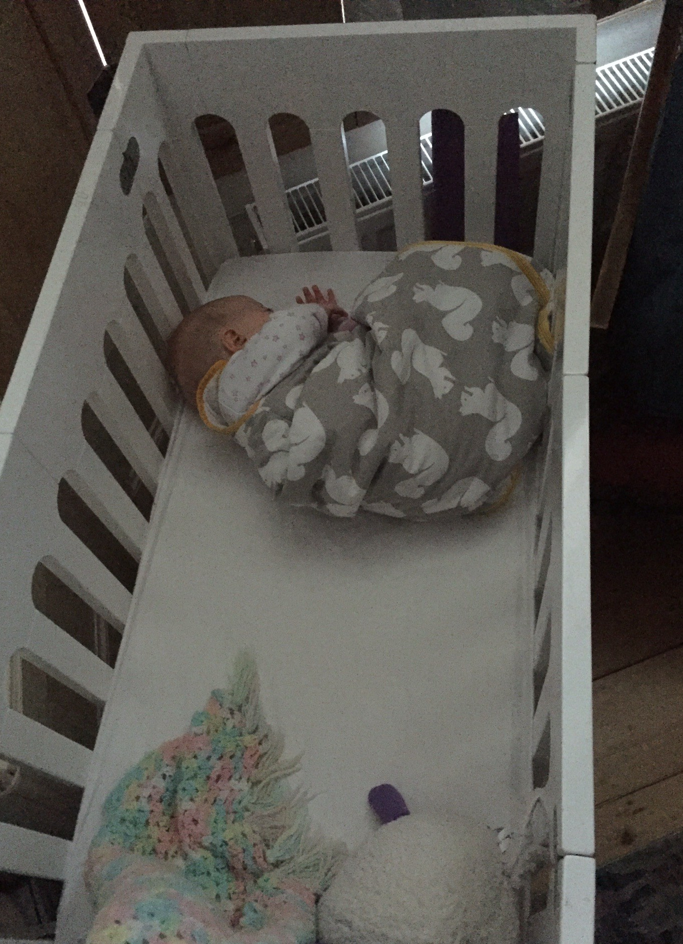 A baby wearing a sleeping bag sleeps curled up in a ball at one end of a cot.