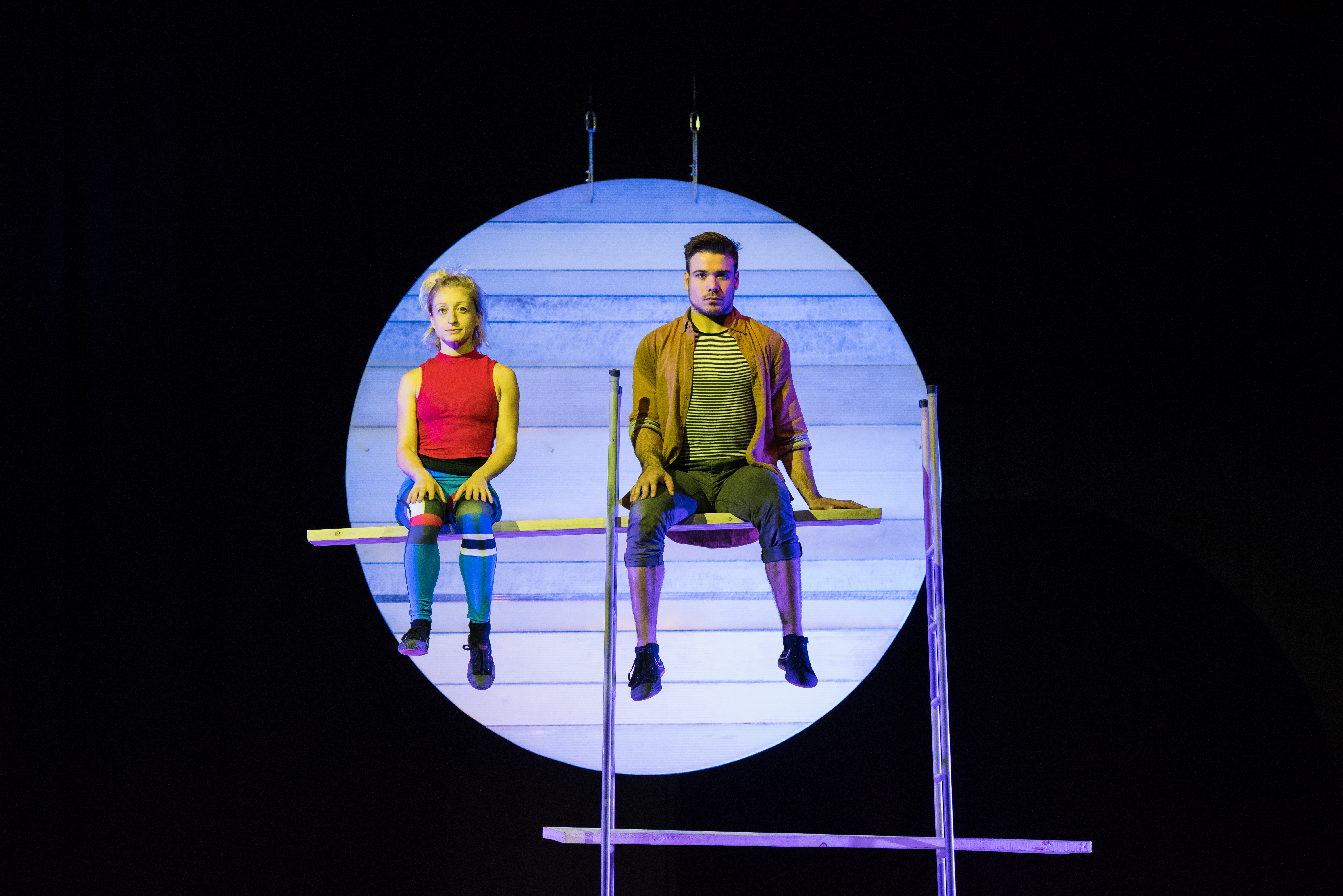 A man and a woman balance on a plank suspended through a ladder, in front of a theatre set of a moon.