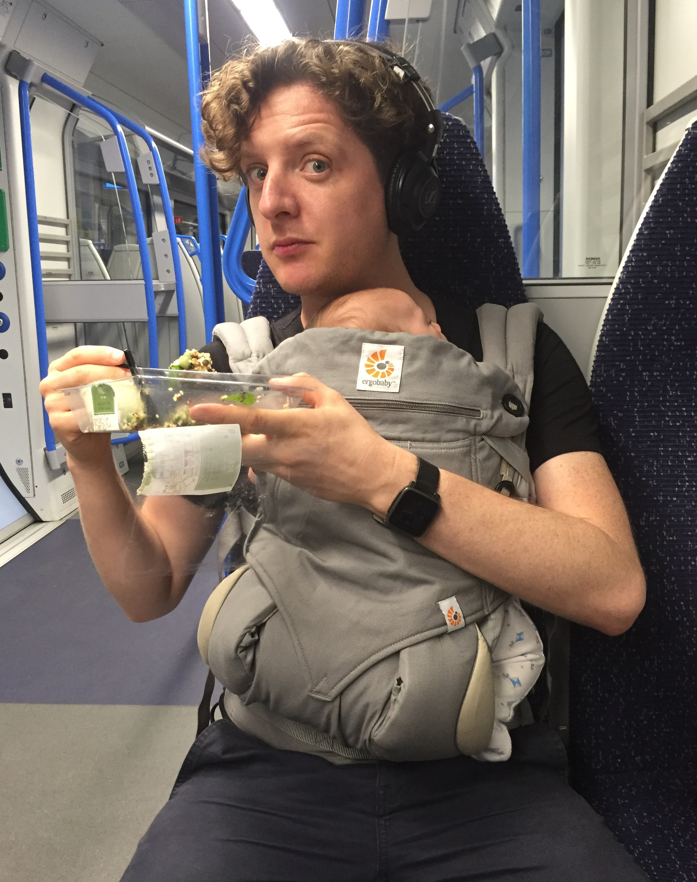 A man with a baby in a sling eats a salad on a train.