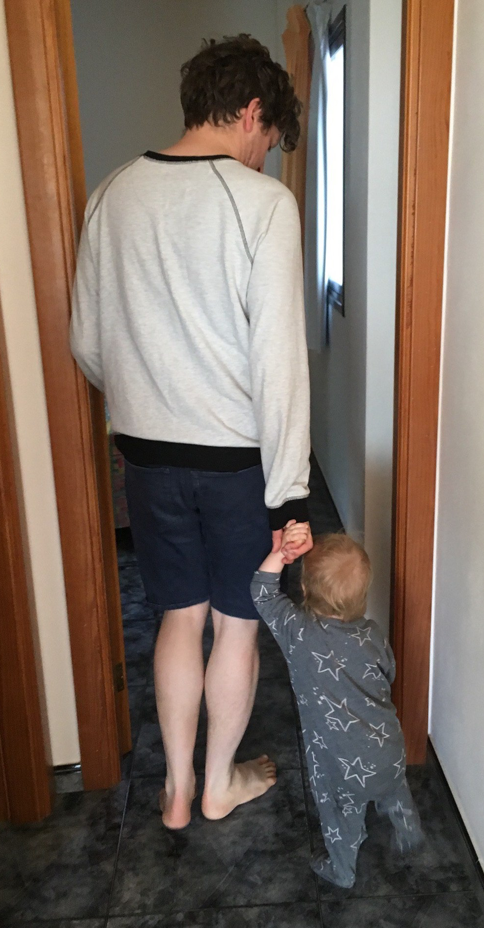 A man and a baby walk down a tiled corridor in an apartment.