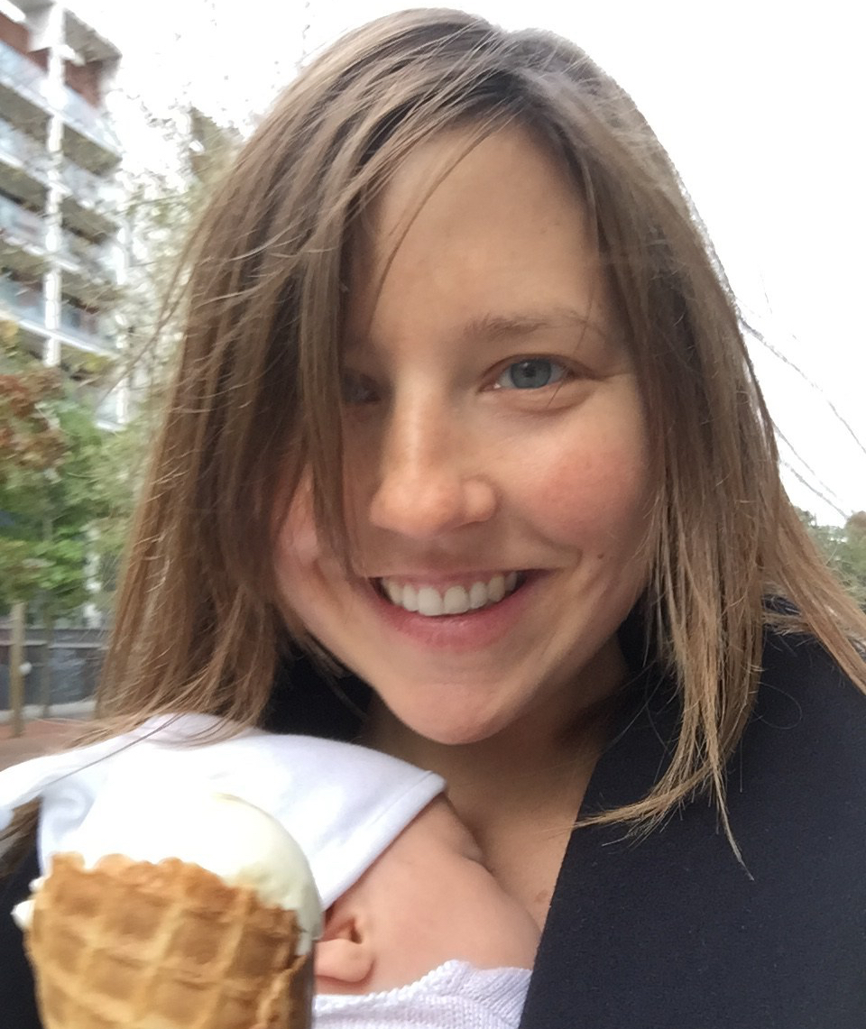 A smiling woman with a baby in a sling holds an ice cream cone.