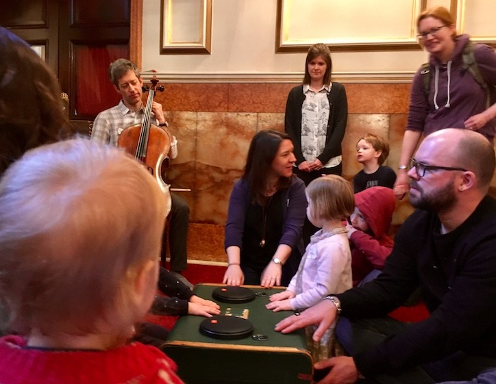 Small children and adults gathered around a suitcase listen to a cello