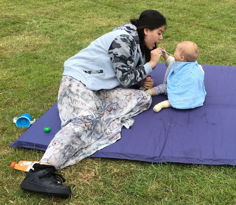 A woman feeds a baby on a blanket on grass