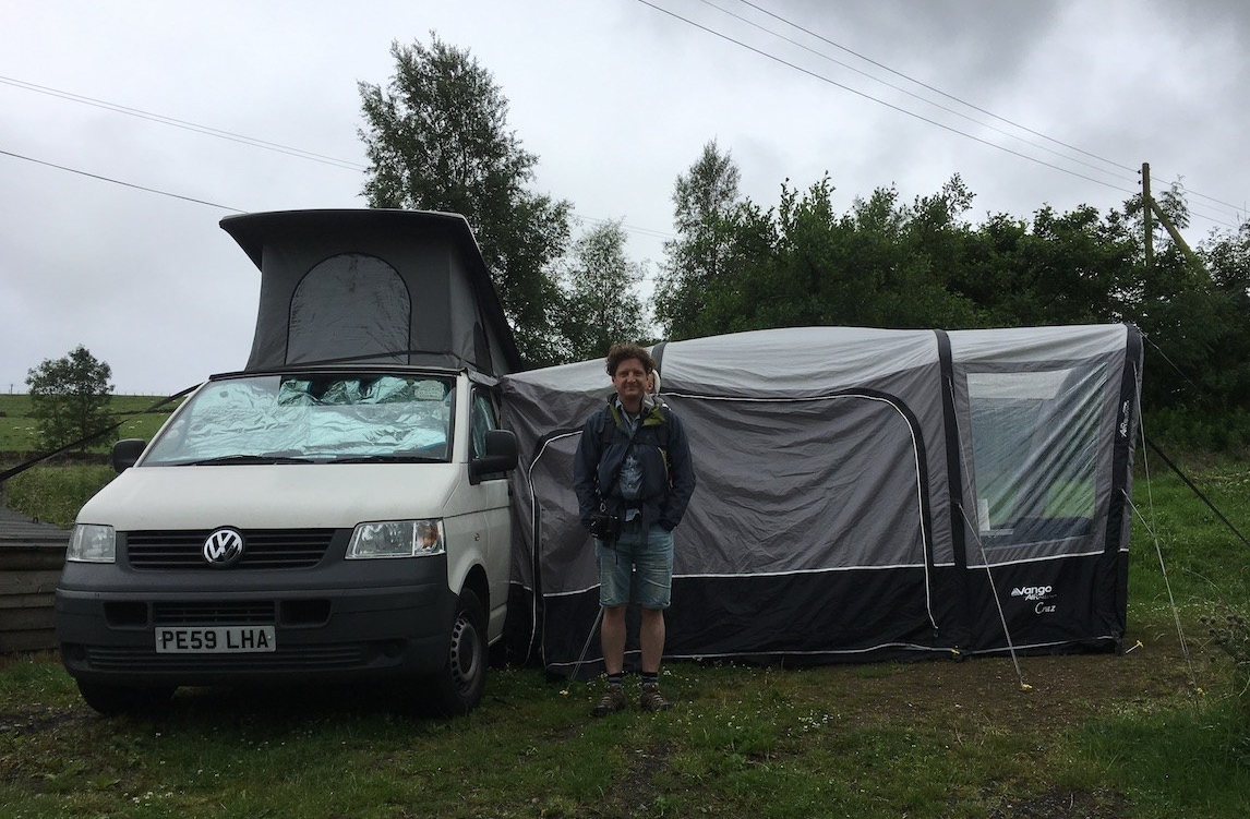 A man with a baby in a back carrier stands in front of a white camper van and awning