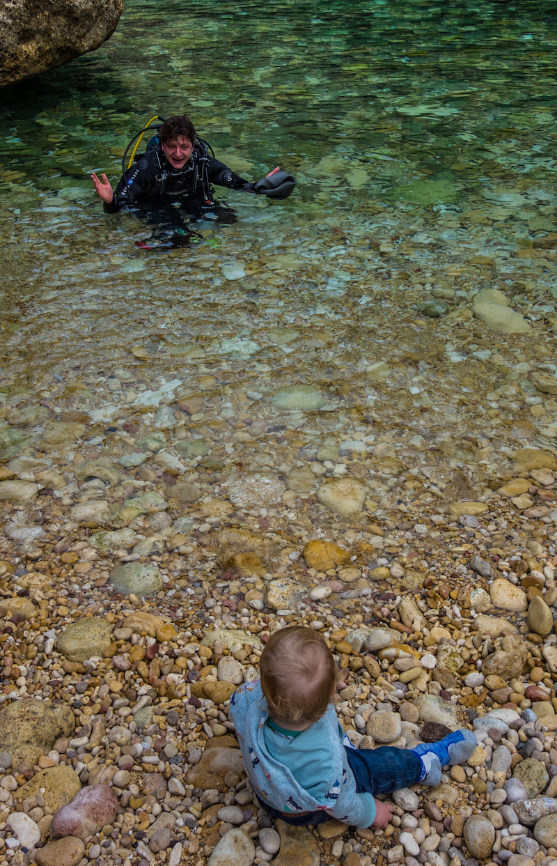 A baby sits on the beach watching a scuba diver come out of the sea