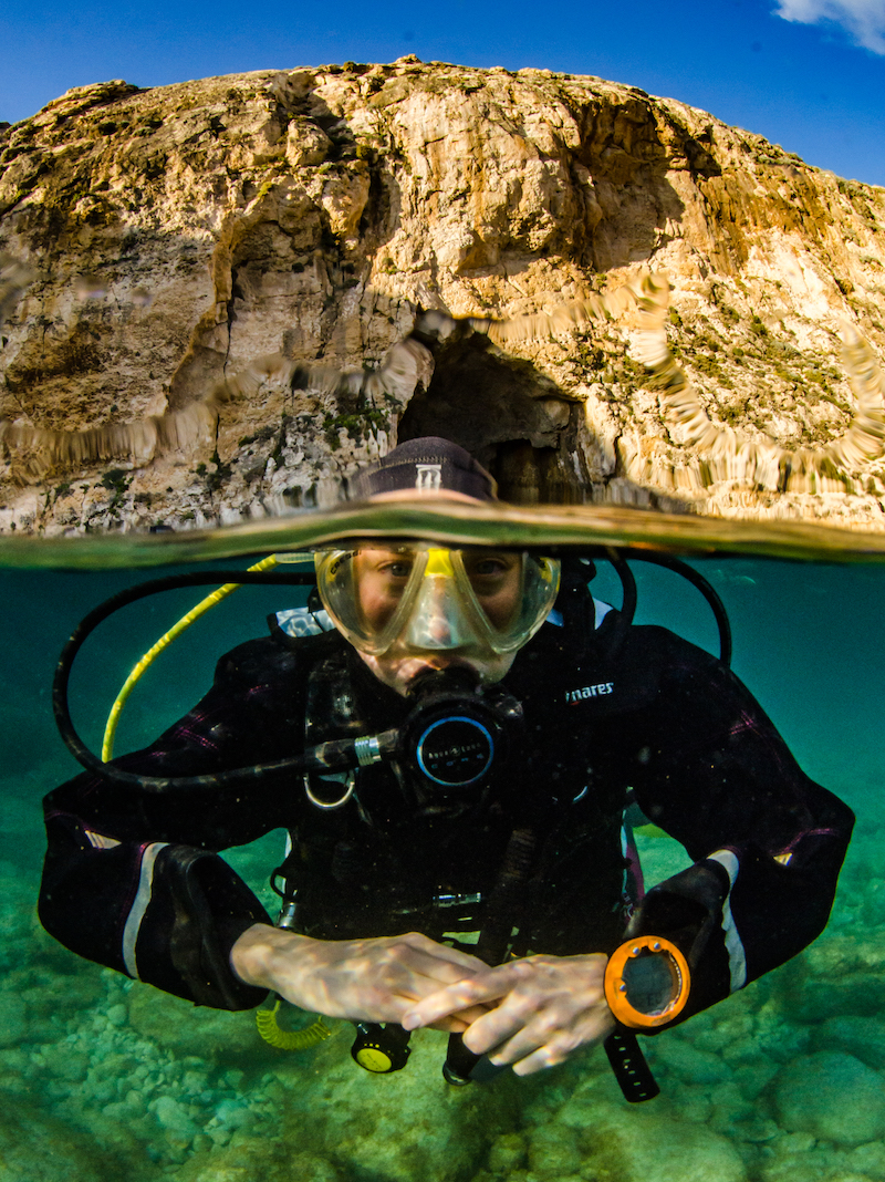 A scuba diver is photographed in front of a channel through a cliff in Gozo