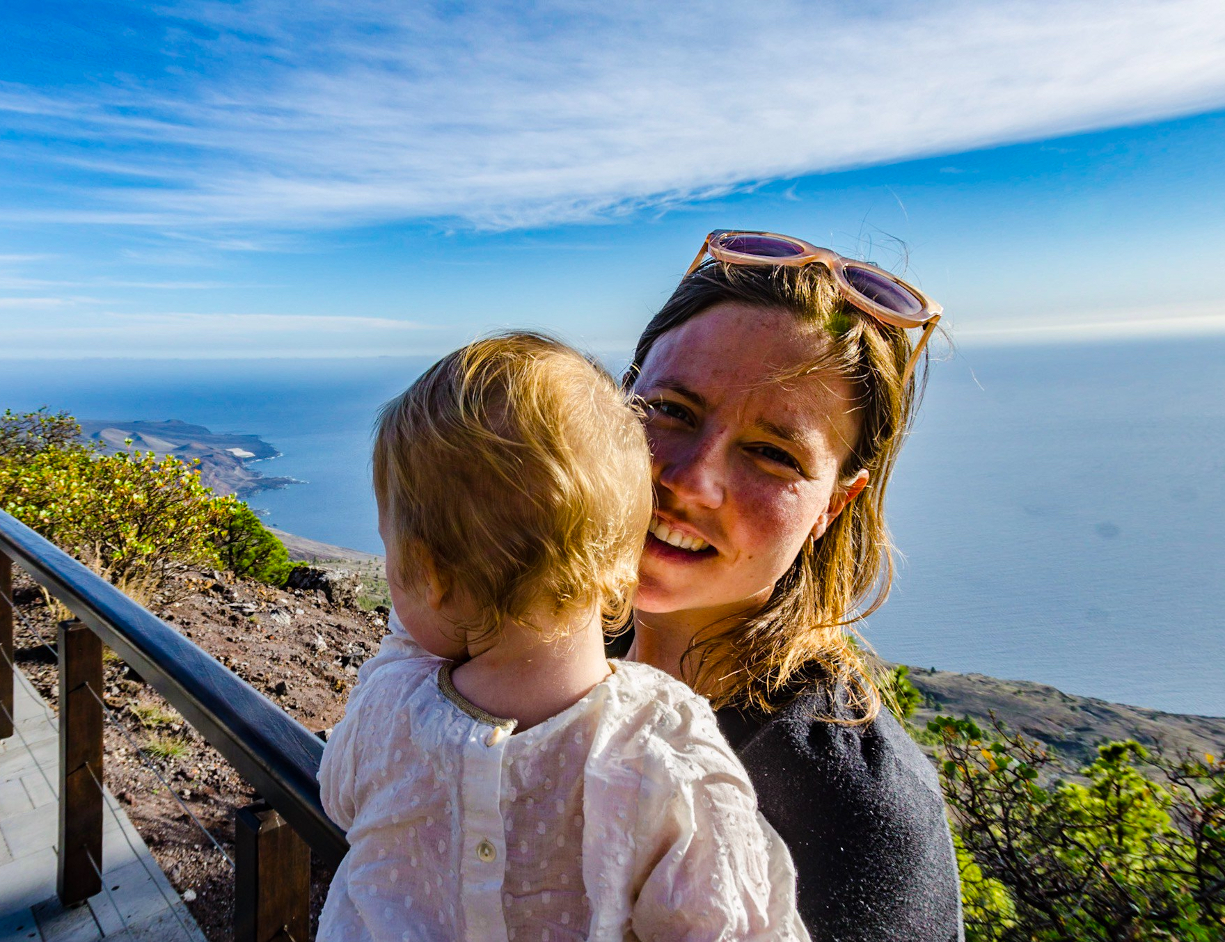 A woman holds a baby in the afternoon sunshine. In the background is the beautiful view of the coastline of El Hierro from the visitor centre at El Julan © Steve Pretty