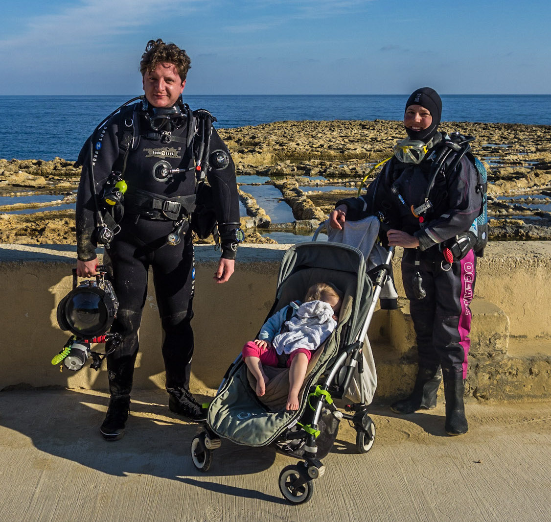 A man and a woman in scuba gear stand at the seaside with their daughter in a pushchair