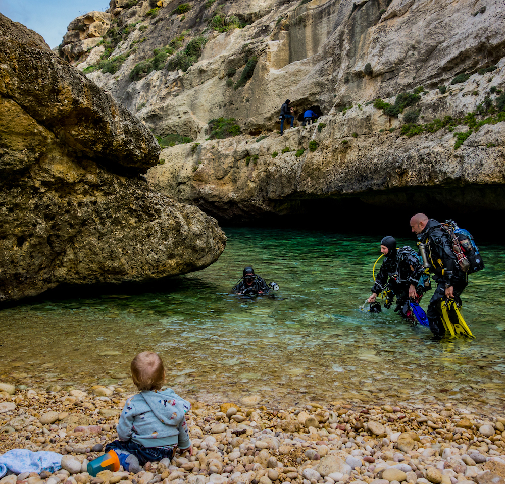 Scuba divers come out of the sea while a baby watches on a beach