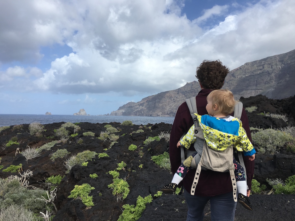 A man hikes through a volcanic seaside landscape with a baby on his back on El Hierro in the Canary Islands