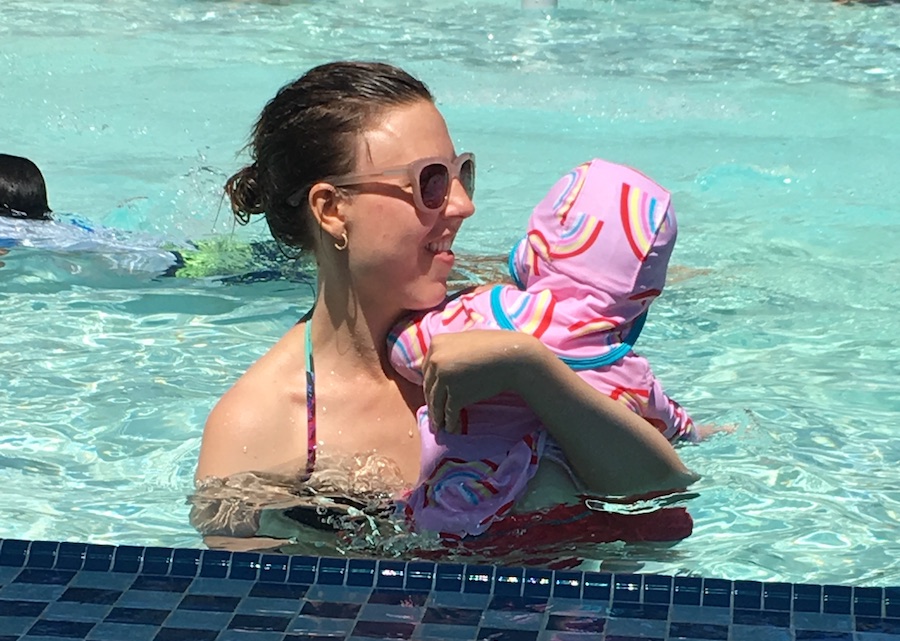 A woman wearing sunglasses swims with a baby at an outdoor pool