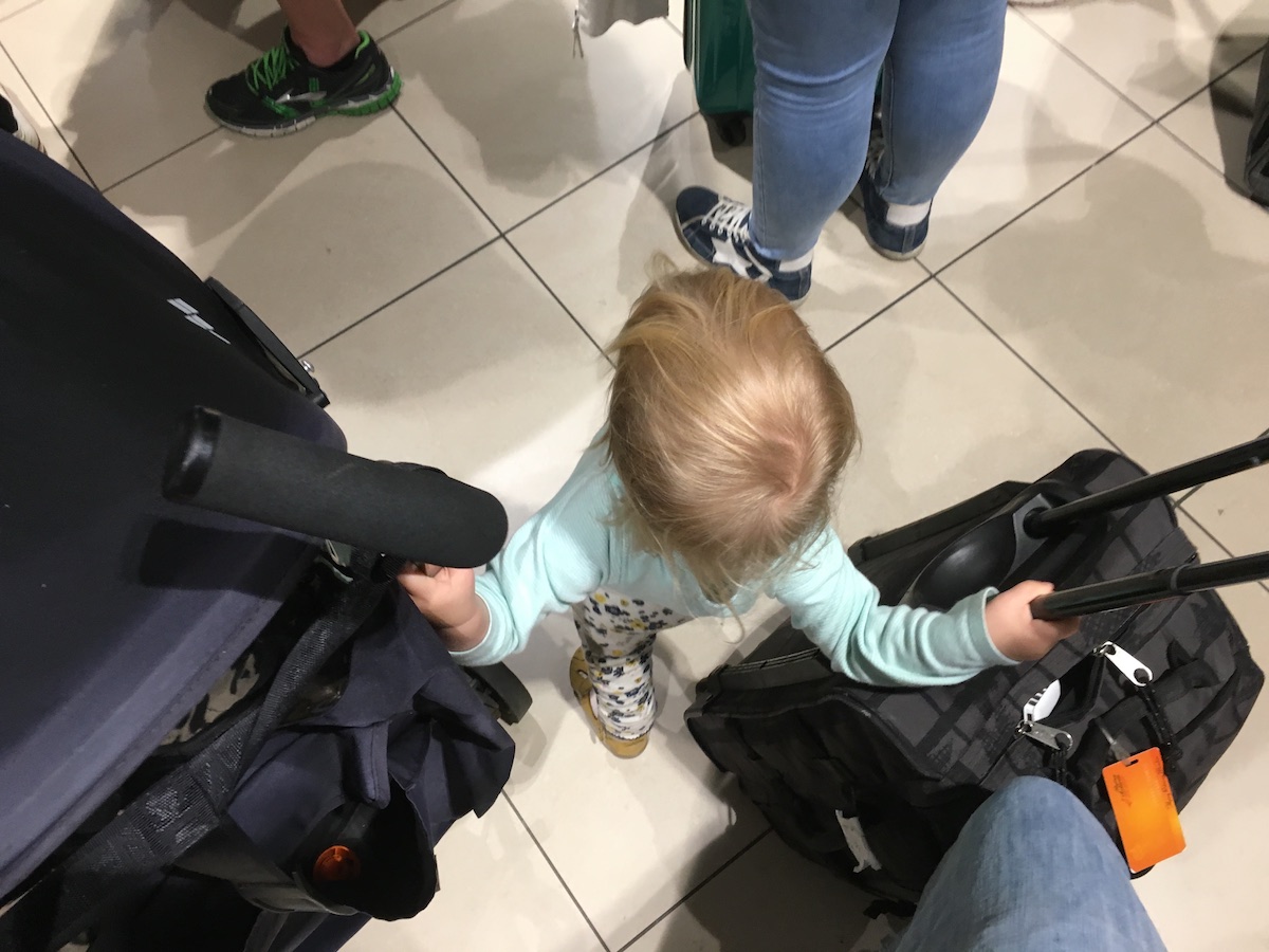 A toddler stands in a queue with a buggy and a wheelie bag