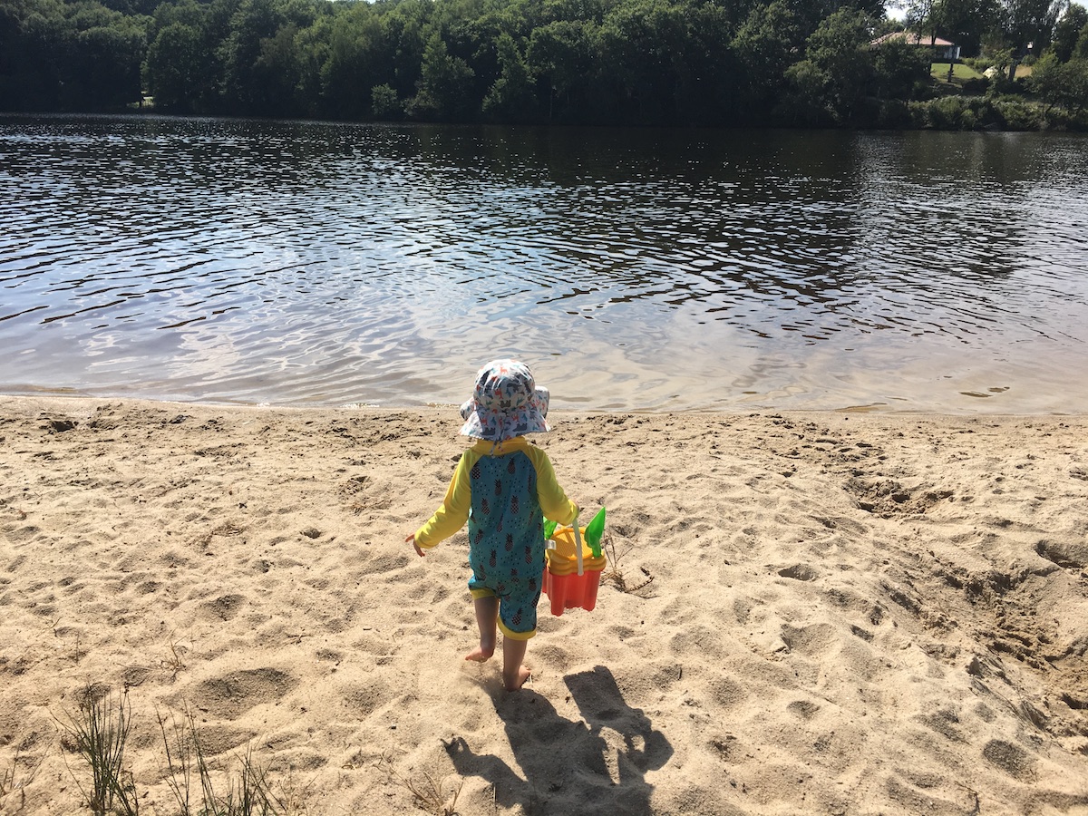 A toddler on a beach at a lake in a 'grow-with-me' sun hat from Twinklebelle and UV rash guard swim vest and shorts from Jan & Jul, the company's other brand